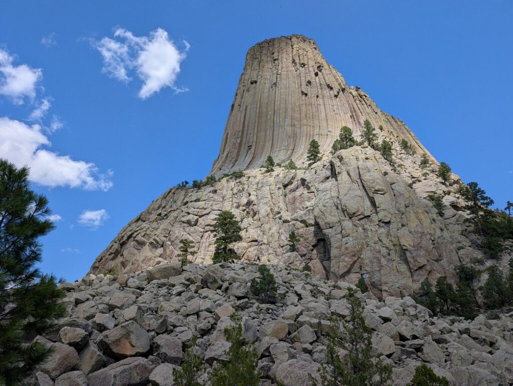 View of Devils Tower in Wyoming from the bottom, looking up. It's a clear blue sky with a few wispy clouds in a perfect blue sky. The monument rises from the ground and reaches to the sky.