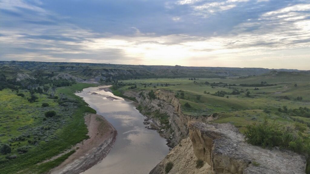 Little Missouri River, North Dakota. River running through the middle of the screen with the North Dakota Badlands on either side. The sun is setting in the west as the river flows towards the sunset in the image.