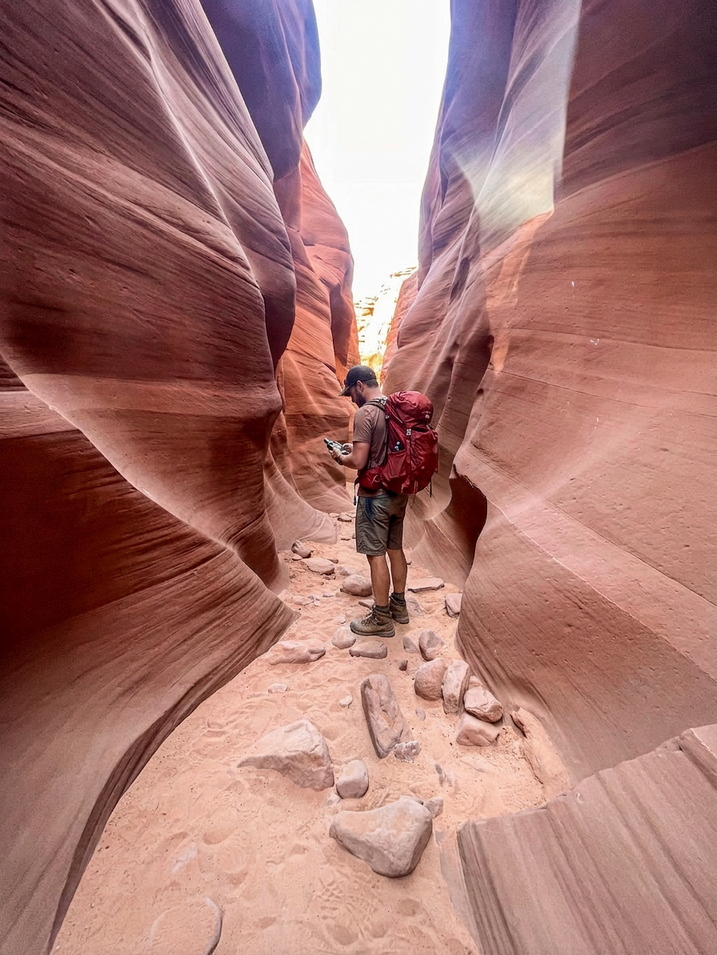 Man looking at handheld GPS receiver while standing in slot canyon. Wearing a red backpack, shorts and a baseball cap.